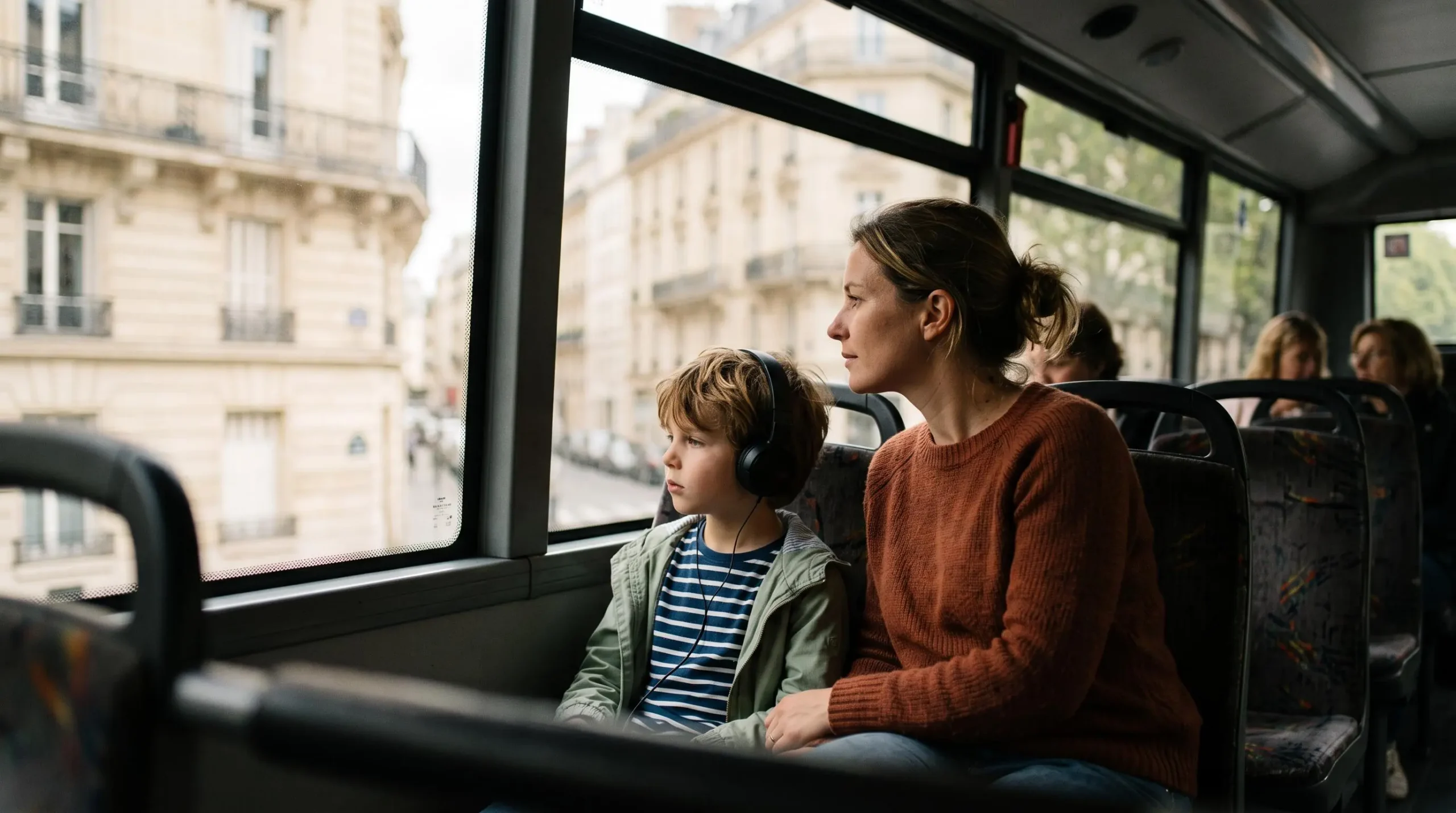 Un enfant portant des écouteurs et son parent regardant Paris par la fenêtre du pont inférieur d'un bus touristique.